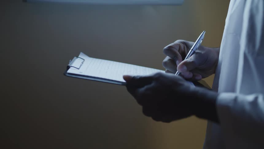 Male doctor holds a clipboard in front of him while standing and using a silver pen to fill out paper work
