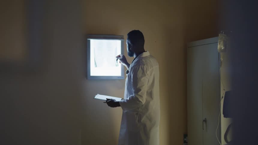 A male doctor stands in front of a lighted x ray board and holding a clipboard