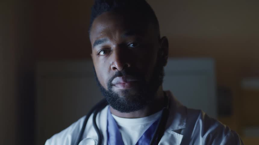 Male doctor with a beard, a lab coat, and a stethoscope around his neck faces the camera with eye contact in the clinic