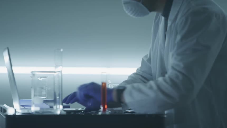 Scientist wearing lab coat and protective gear takes notes from a laptop on a desk