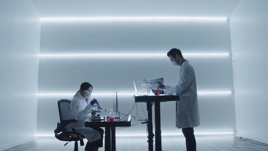 A female scientist types on a computer and uses a microscope while a male scientist examines an xray and writes on a clipboard across from her