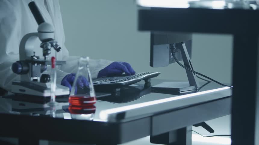 A medical person sits at a desk with a microscope beaker computer and keyboard