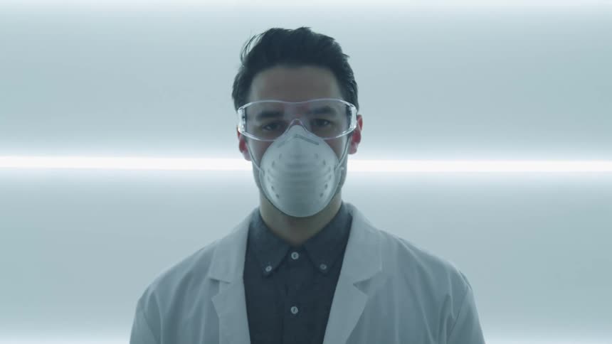 A male scientist in a lab coat safety goggles and dust mask raises his head to look into the camera