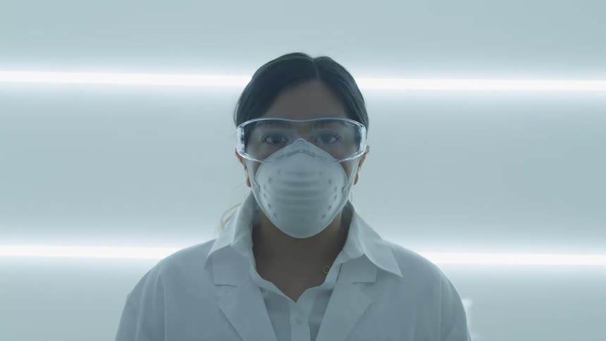A female scientist in safety goggles and a dust mask slowly raises her head to look into the camera