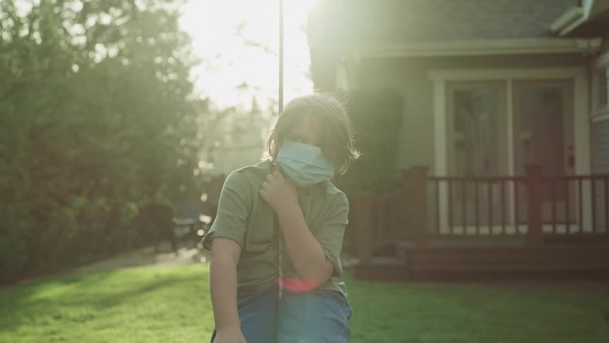 Boy wearing a face mask sitting on a swing in the yard of a house at sunset