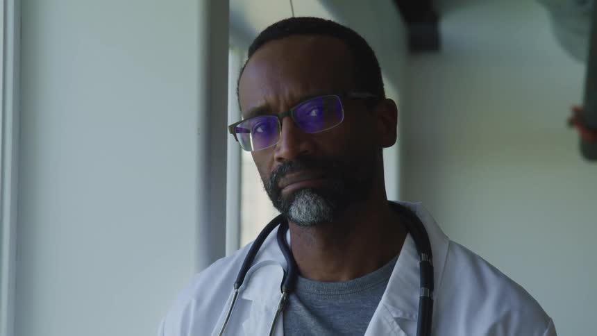 A doctor wearing glasses and a stethoscope stands in a room near windows