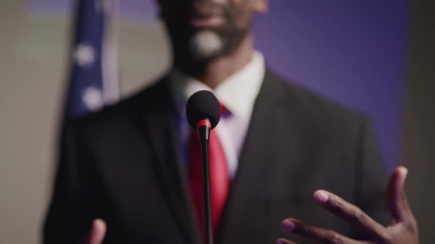 A man in a suit talks while standing at a microphone in front of an american flag