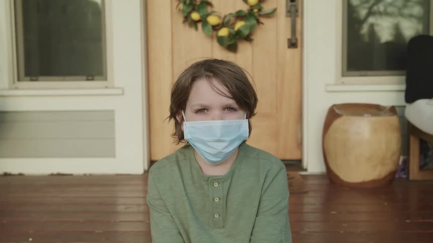 Boy with a long sleeve green t shirt and a light blue mask kneels down on wooden flooring and faces the camera with eye contact