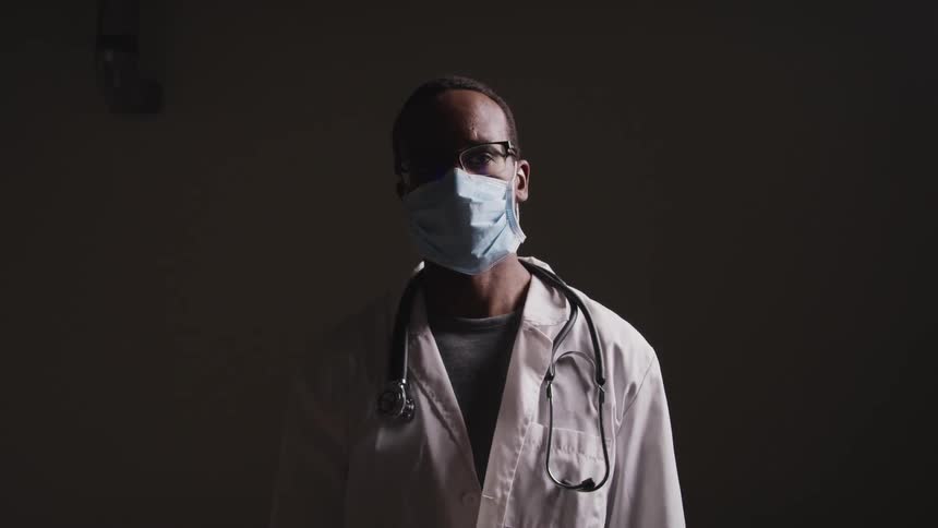 Close up of a doctor wearing a face mask with stethoscope over his shoulders in a dimly lit room