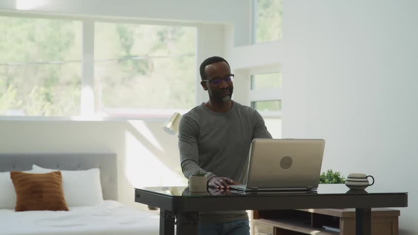 Man wearing glasses uses a laptop in an stand up desk on the bedroom
