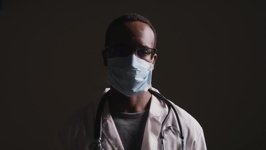 Black male doctor with a lab coat glasses a stethoscope and a blue mask raises his head and faces the camera with eye contact on a dark background