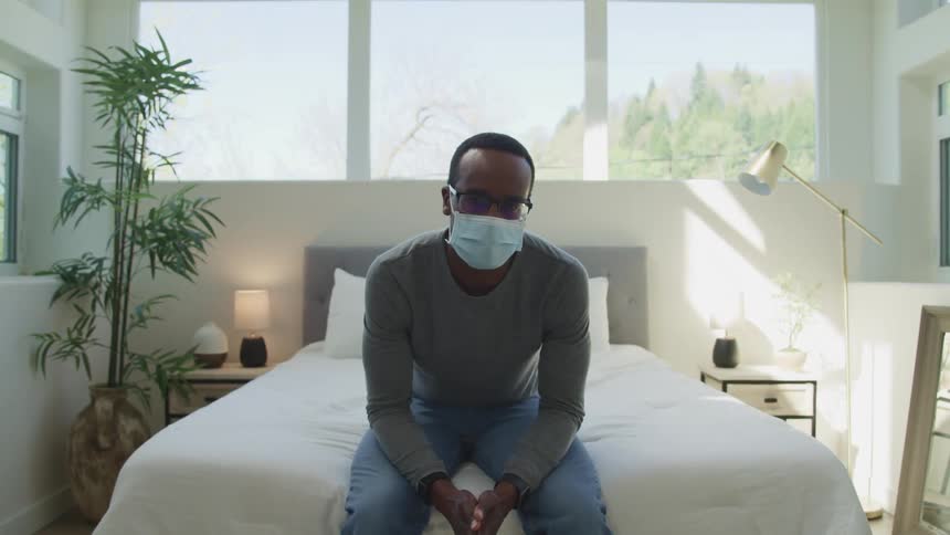 A man sits on his bed with a mask on