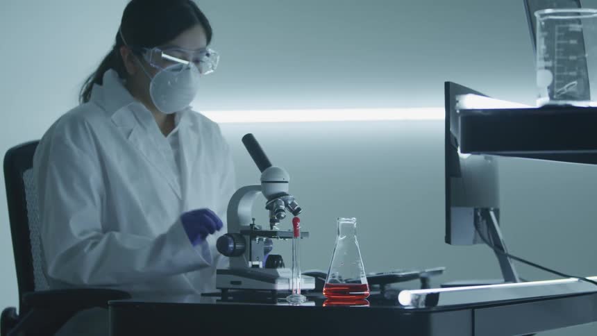 A woman with goggles a mask gloves and lab coat looking into a microscope