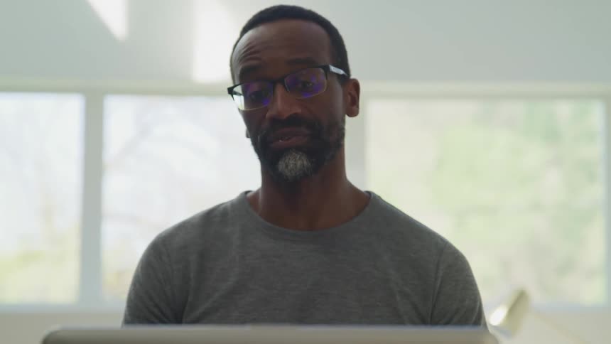 Bearded man wearing glasses talks on a laptop video call in a bedroom at home