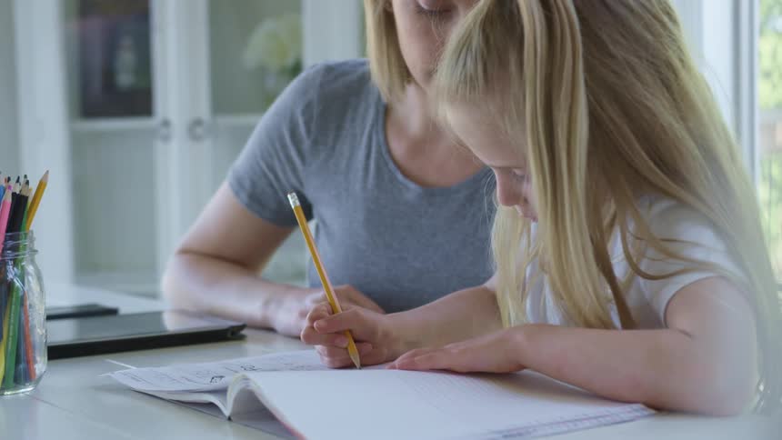 A woman sits at a table with a little girl and watches as she writes in a notebook