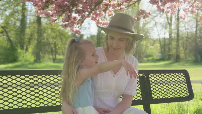 A woman embraces a young girl as they sit on a park bench and the girl waves her hand at something out of frame