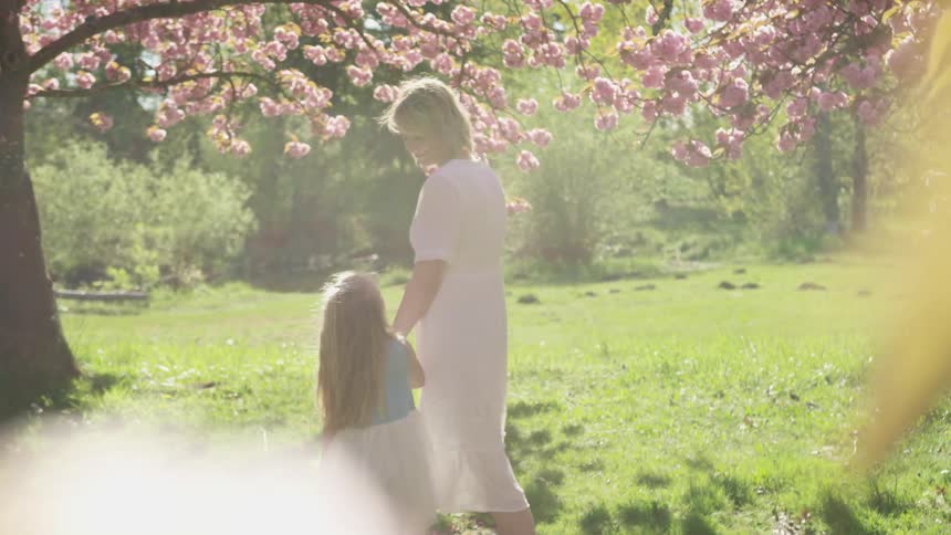 A woman holds her daughters hand as they walk under a cherry blossom tree