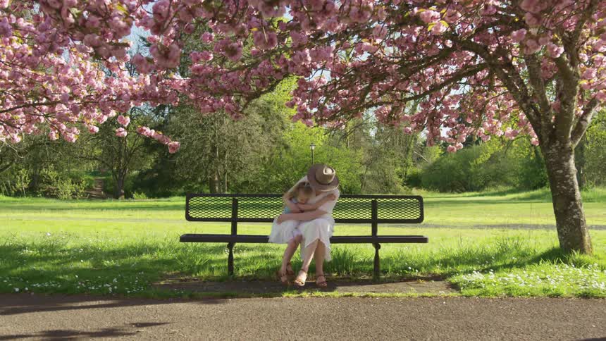 Mother and daughter playfully hug each other in the bench of a park under a cherry blossom tree