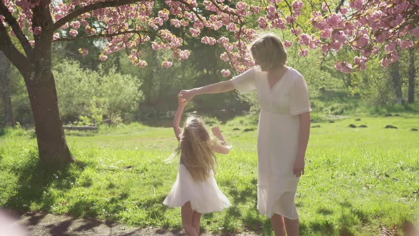 A little girl holds her moms hand and twirls around under trees filled with flowers