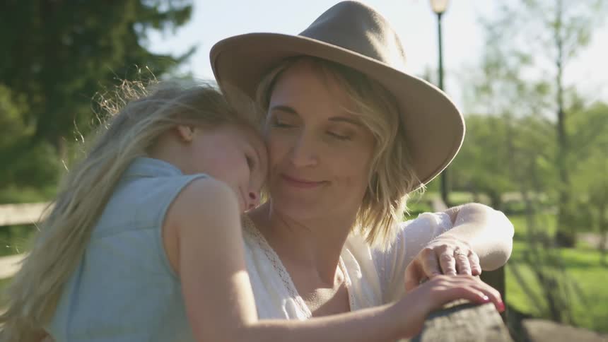 A woman in a sunhat holds and smiles at her young daughter as she leans against a wooden fence on a sunny day