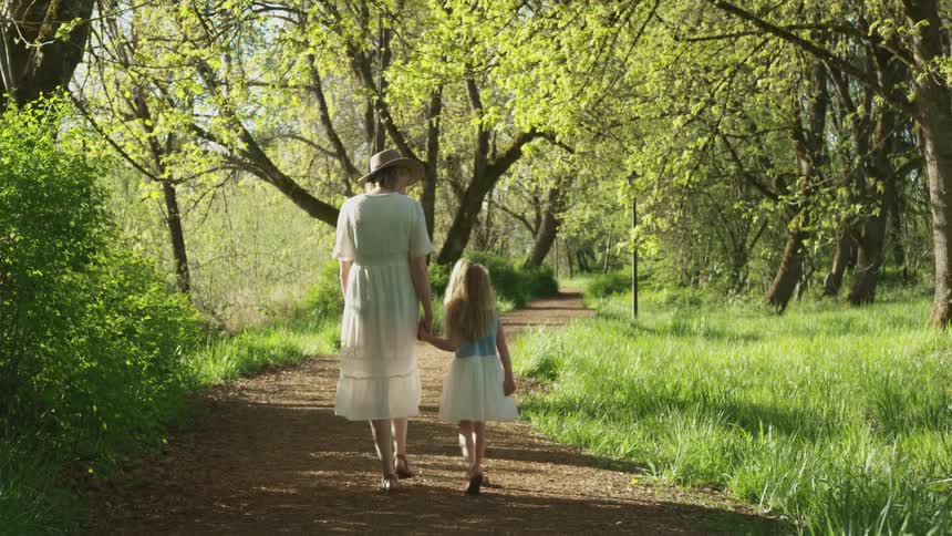 A woman holds her daughters hand as they walk down a trail in a park while wearing dresses