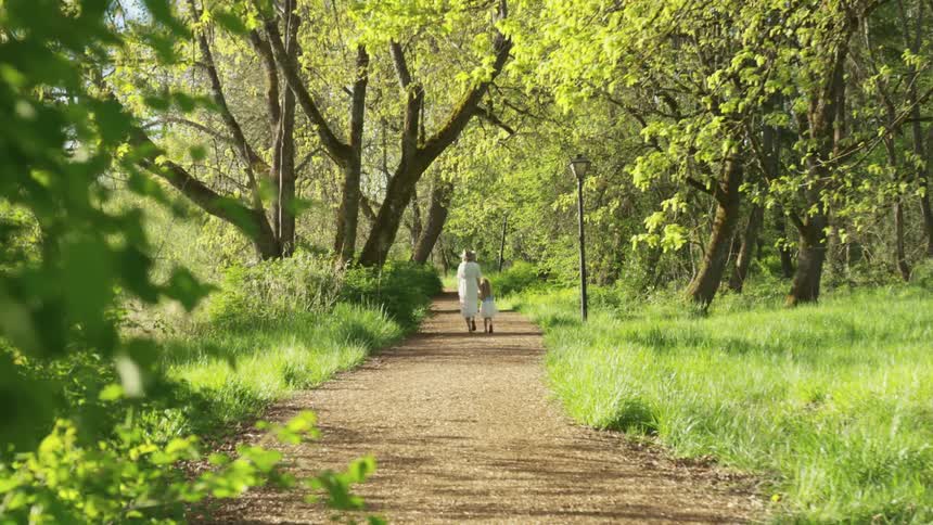 A woman holds her young daughters hand as they walk down a wide dirt trail in a park