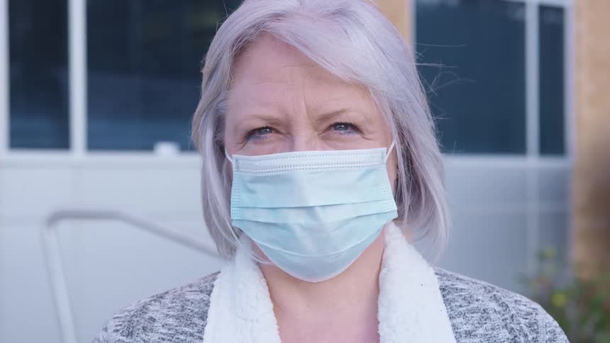 Elderly woman with grey hair and a blue mask faces the camera with eye contact