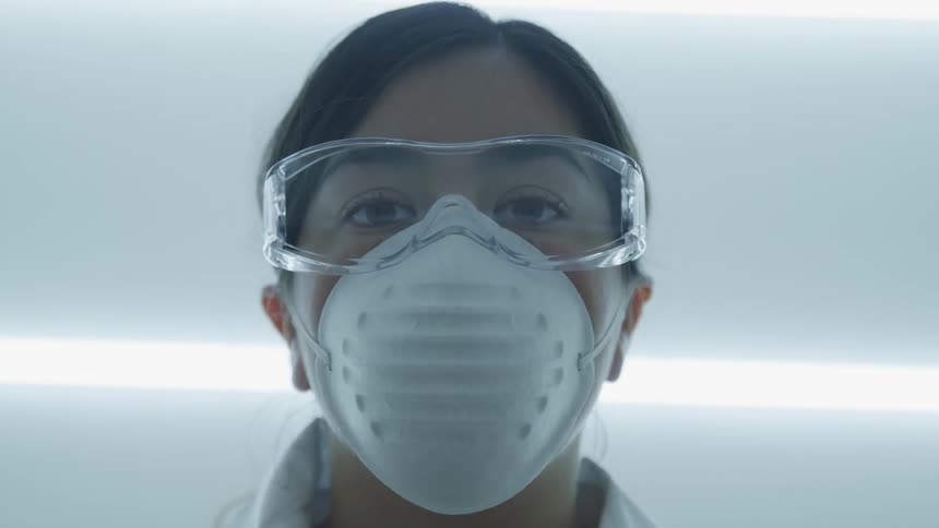 A young female scientist wearing safety goggles and a dust mask slowly raises her head to look into the camera