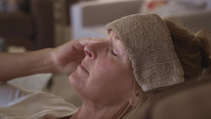 A woman laying down with her eyes closed places a damp towel on her forehead
