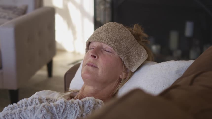 A woman sleeps on a sofa with a folded towel resting on her forehead
