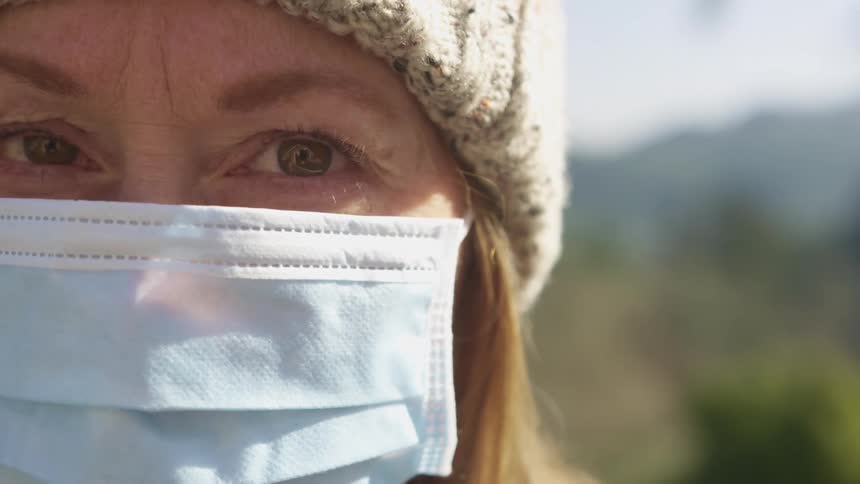 A woman wearing a knit hat and a surgical face mask looks directly towards the camera