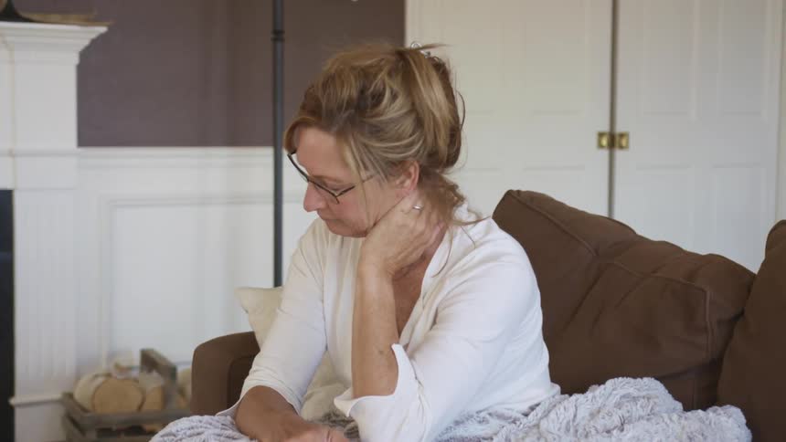 Middle aged blonde woman with glasses and a white shirt sits on a brown couch and rubs her neck in the living room at home
