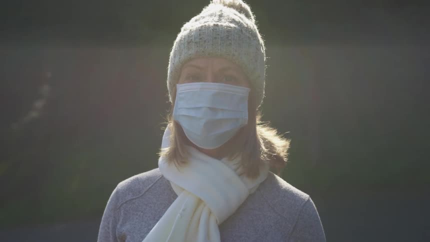 Woman with a grey beanie a white scarf around her neck and a light blue mask faces the camera with eye contact