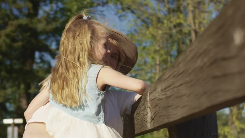 Mother holds daughter in her hands while reclining in a wooden fence near the woods