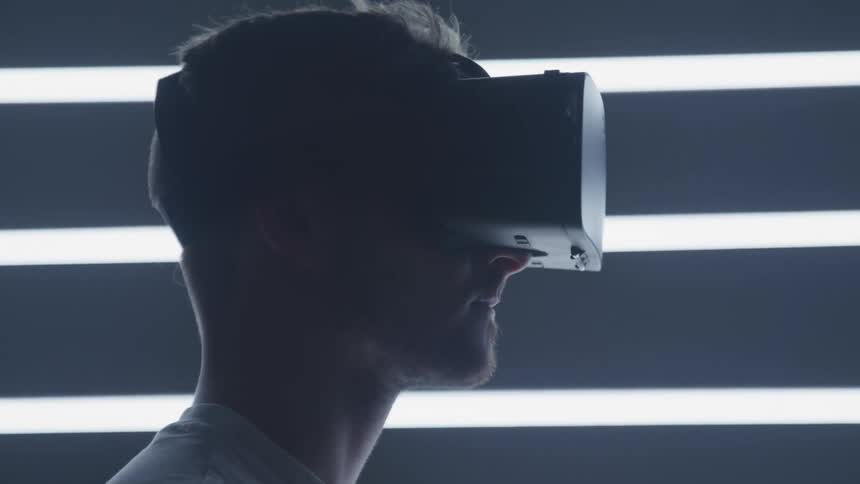 A man in a virtual reality headset in a room with fluorescent lights on the wall slowly raises his head