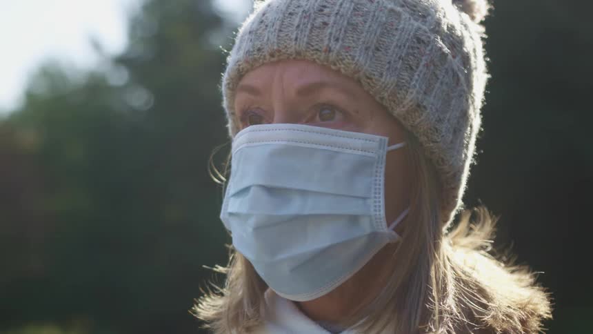 Middle aged blonde woman with a grey beanie and a light blue mask looks into space