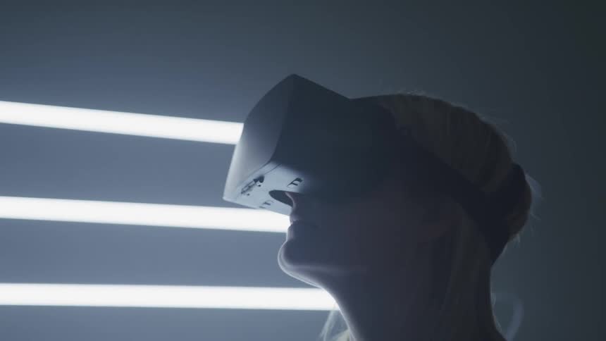 A woman wearing a virtual reality headset stands in front of wall where a row of fluorescent lights are mounted