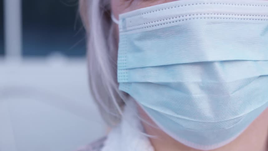 Elderly woman with grey hair and wearing a light blue mask faces the camera