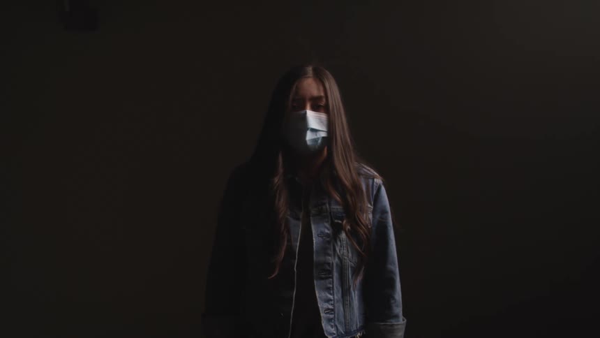 Young woman wearing a face mask looks at the camera while standing in a dimly lit room