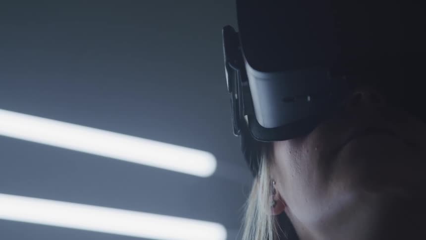 A woman in a virtual reality headset turns her head while standing near a wall wear fluorescent lights are mounted