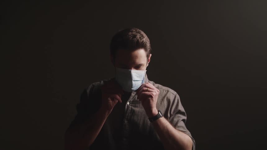 Man with a brown shirt and wristwatch wears a light mask on a dark background