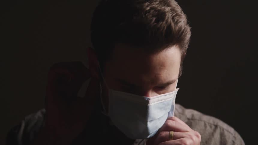 Man with a grey shirt wears a light blue mask and faces the camera with eye contact on a dark background