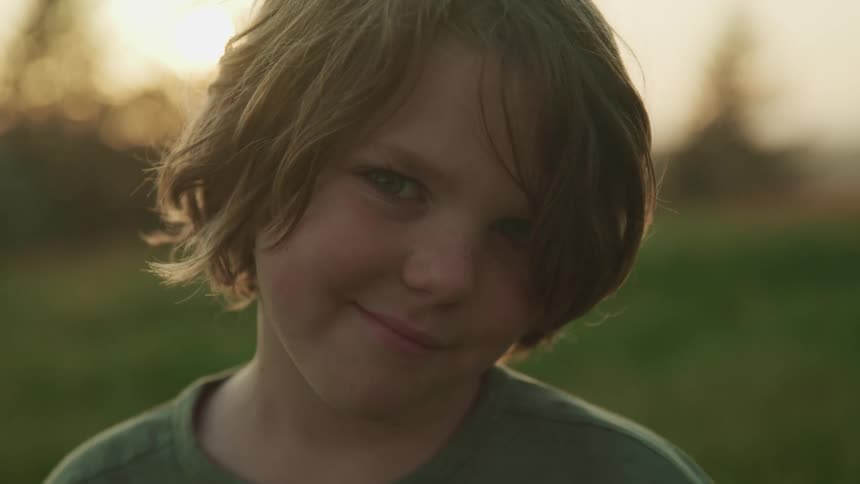 Boy with long hair and a grey t shirt stands on green grass and faces the camera with eye contact at sunset