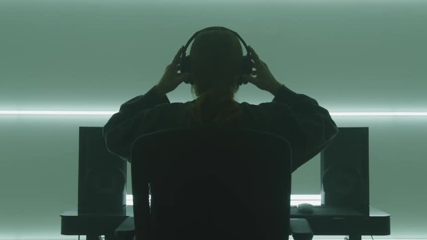 A young woman slowly places a pair of headphones over her ears as she sits at a computer