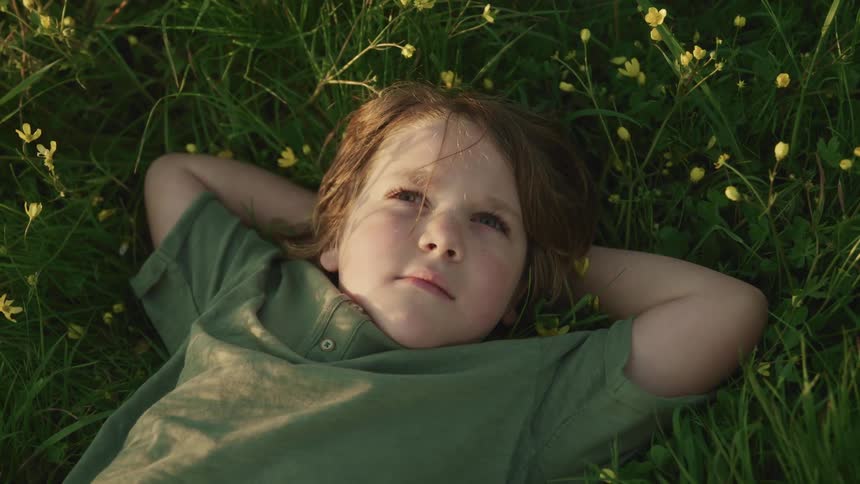 Little boy with long hair and a green t shirt lies down on the green grass