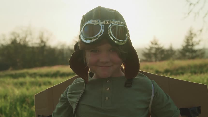 Small boy wearing a vintage aviator hat with goggles and cardboard wings stands in the grass at sunset