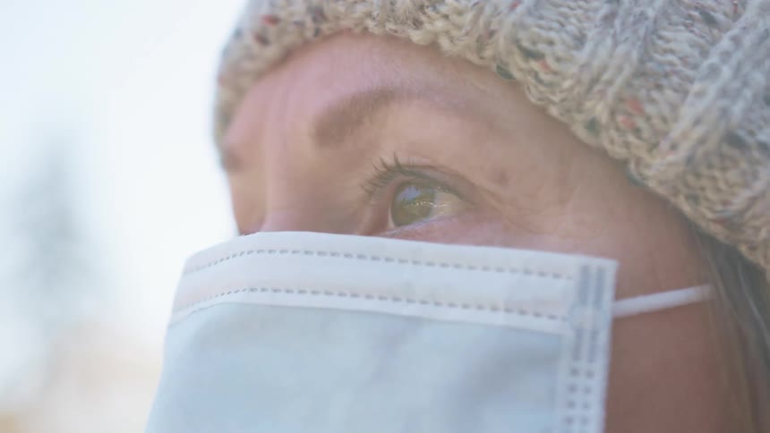A woman in a medical mask stands outdoors during the day and gazes upwards