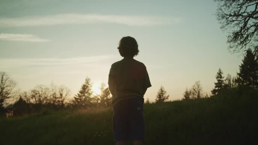 A young boy stands outside and looks towards the setting sun