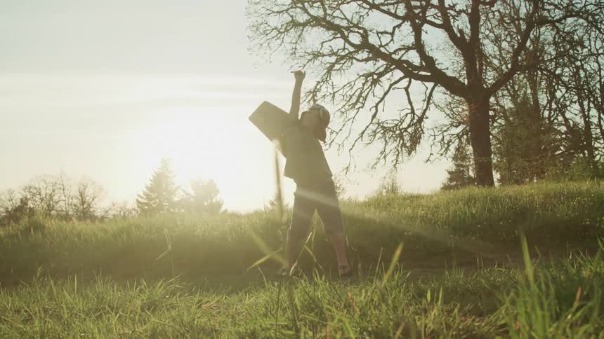 Little boy wearing vintage aviator hat with goggles and cardboard wings plays outside at sunset