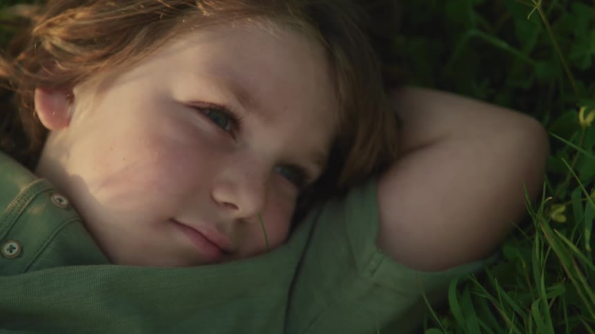 Boy with long hair and a green shirt lies down on green grass